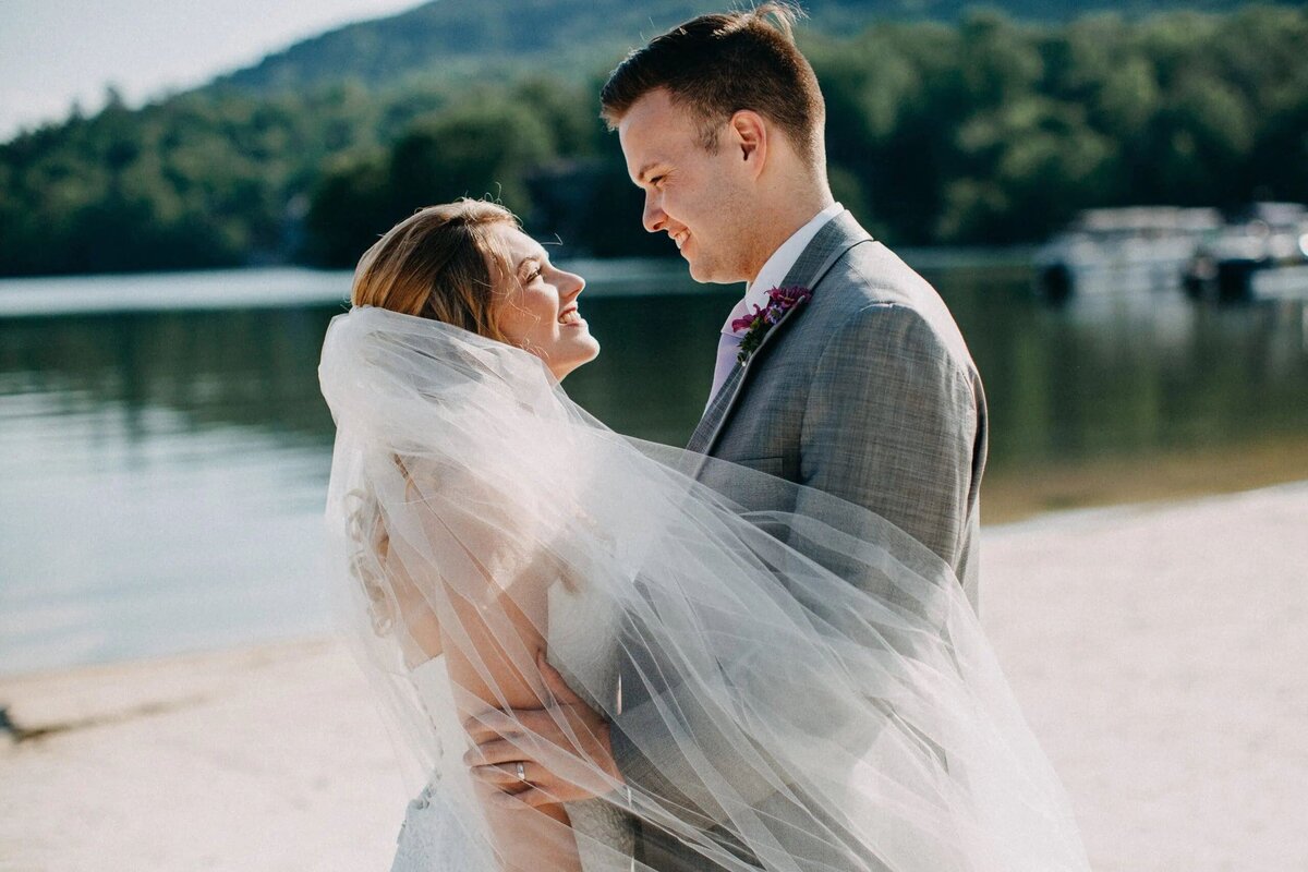 A bride and groom share a close embrace on a lake shore, with the bride’s veil softly blowing in the wind, surrounded by natural beauty.
