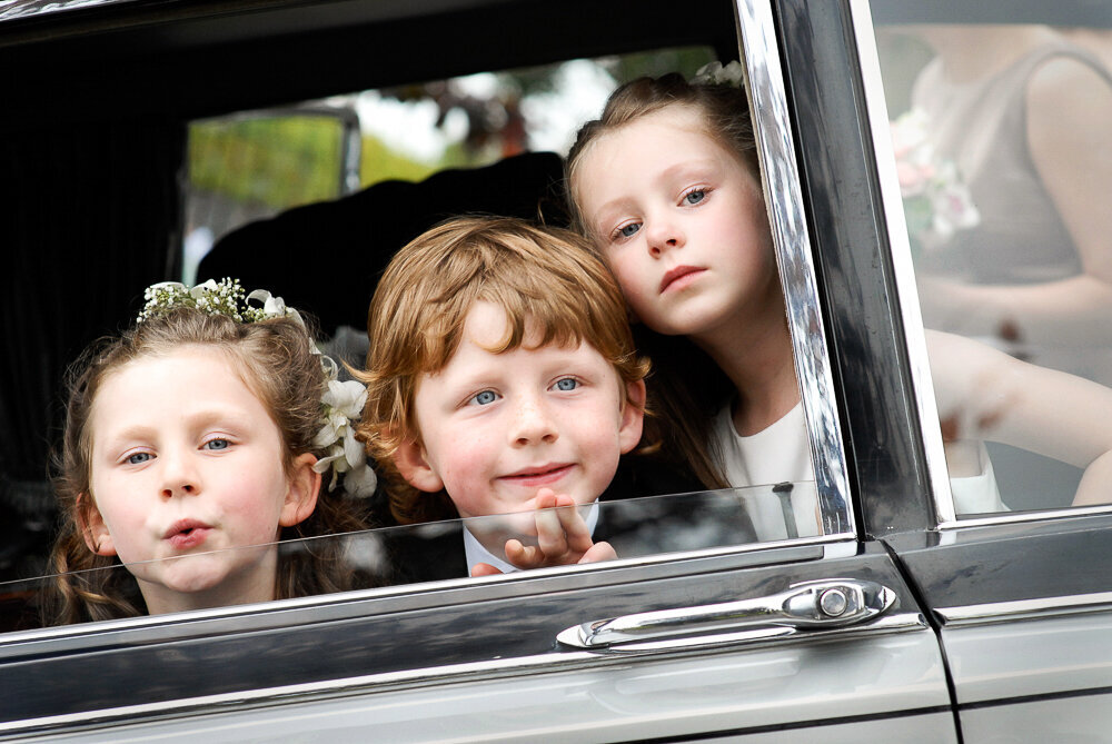 two flower girls and one page boy looking out the window of a vintage wedding car