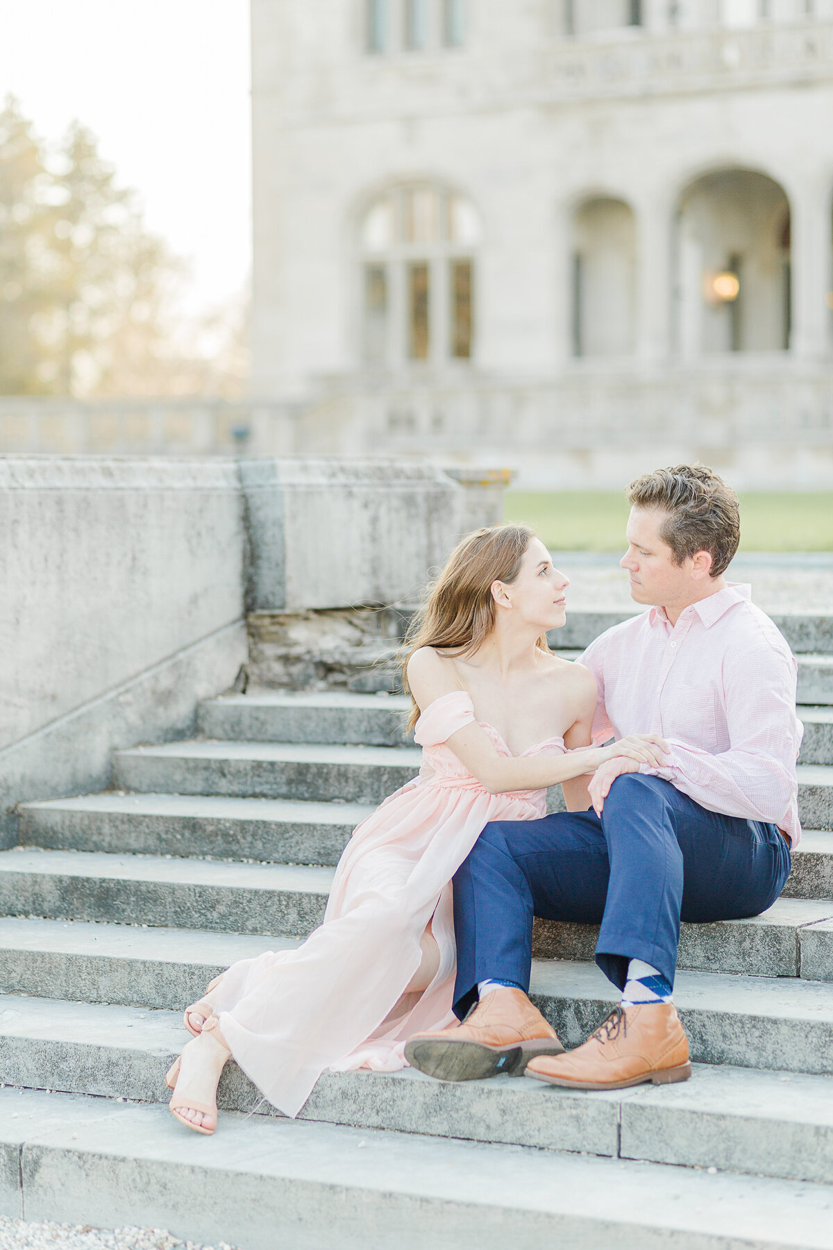 Man and woman sit on the stone steps at Salve Regina. The man is on the step higher than the woman. She is leaning on his lap and looking over at him. The stone buildings are featured prominently in the background. Captured by best RI wedding photographer Lia Rose Weddings