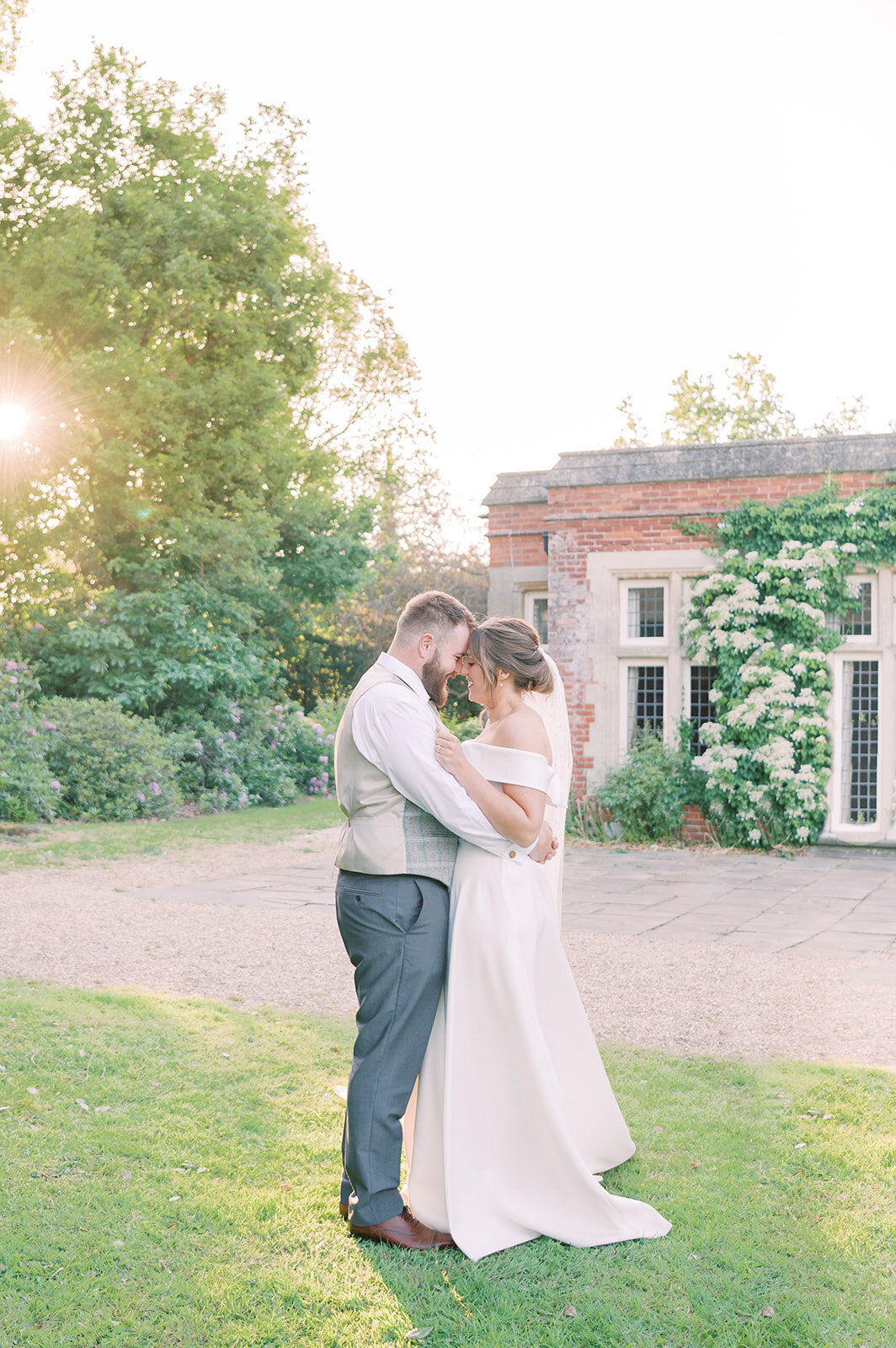 Bride and groom are cuddling face to face with their foreheads touching, the bride is wearing a white dress and the groom is in a suit . The sunset is shining through the trees to the left of the couple