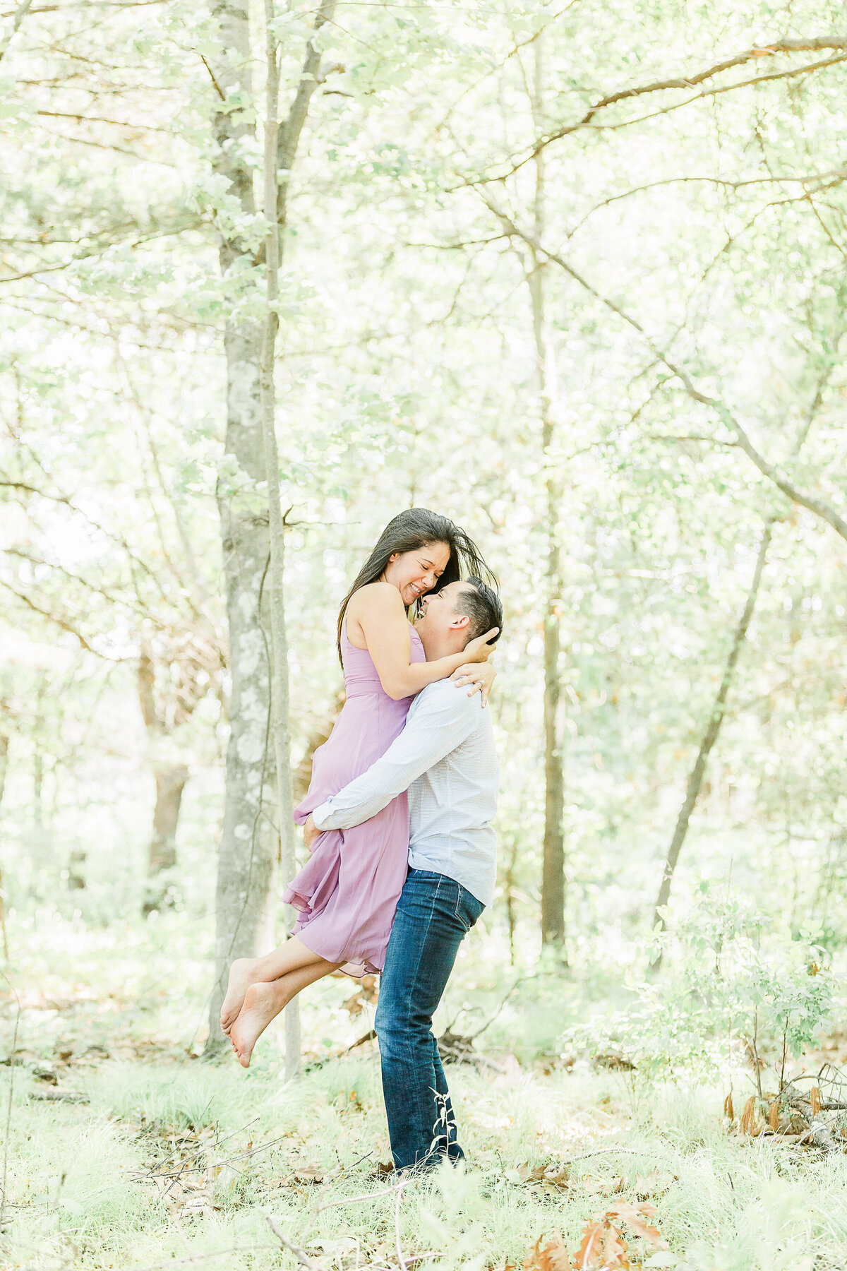 A man picks up his fiance at their Reading Town Forest engagement photoshoot. The woman's arms are wrapped around the groom's neck and their faces are close together and looking at each other. Captured by best MA wedding photographer Lia Rose Weddings.
