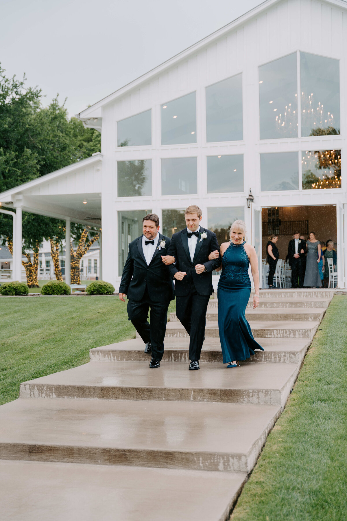 A young man in a tuxedo walks arm-in-arm with an older man and woman down steps from a white farmhouse-style venue, conveying a formal and joyous occasion.