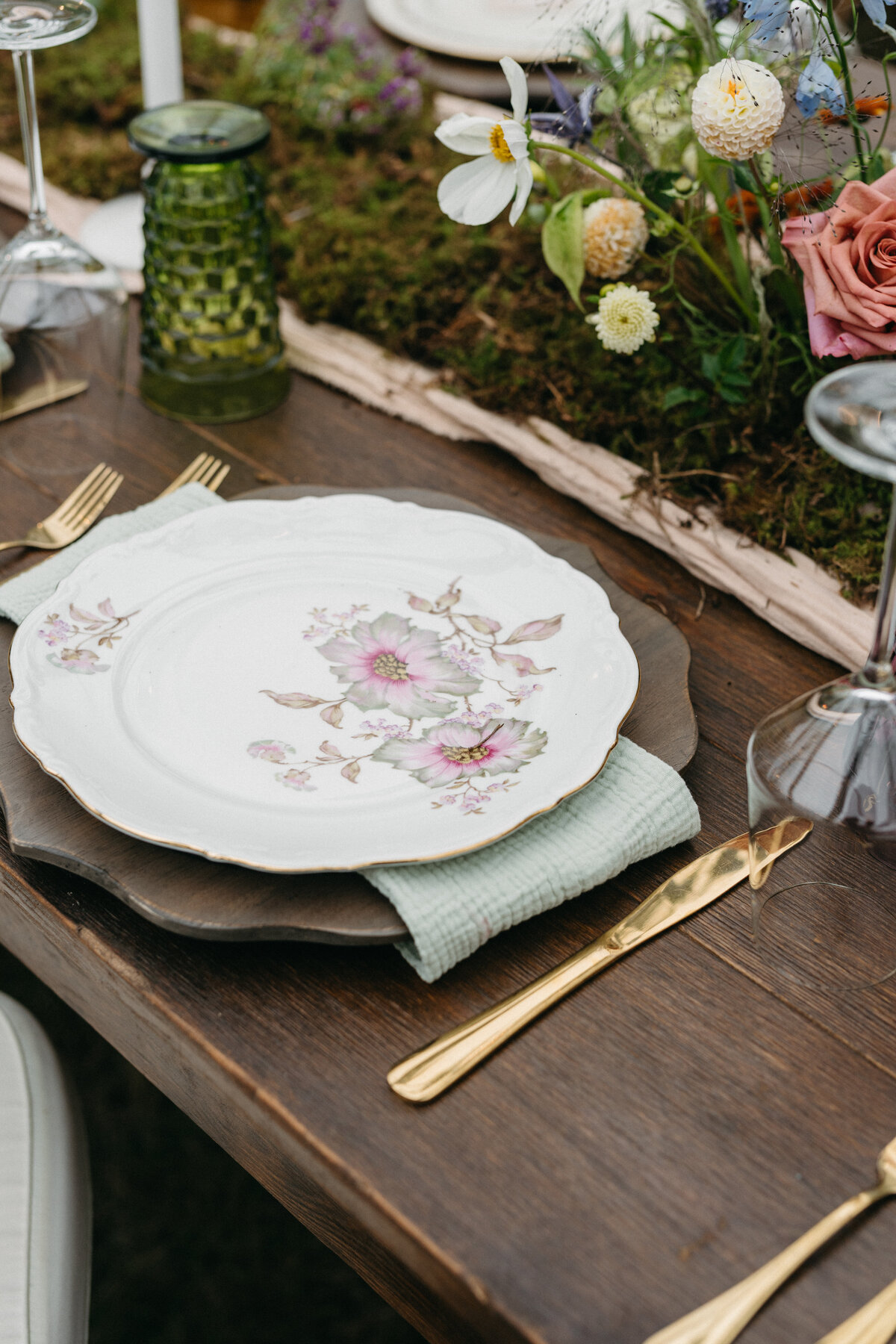 Elegant wedding reception place setting featuring a vintage floral china plate, soft green napkin, gold flatware, and a moss and wildflower centerpiece runner at The Nest in Northwest Arkansas.