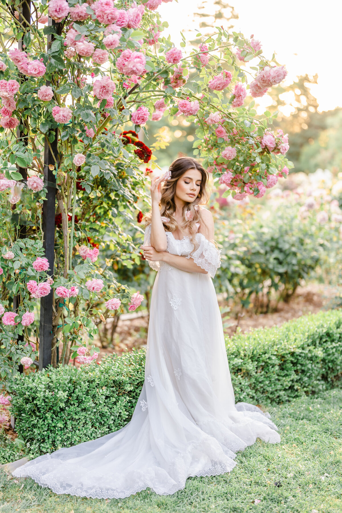 Woman stands dressed in a long white gown in a rose garden with floating rose pieces in her long hair with her eyes closed and hand gently touching a flower piece photographed by Bay area photographer, Light Livin Photography.