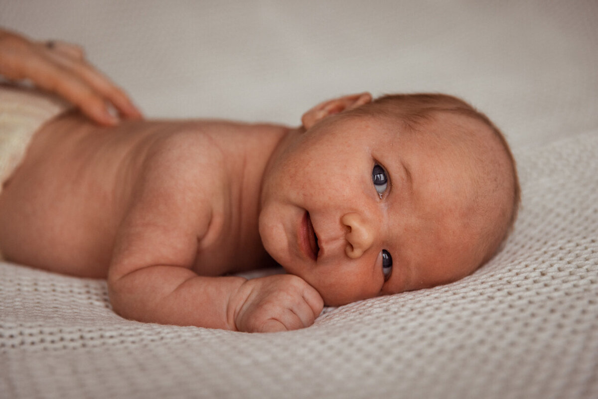 Livsstilsfoto portrett av baby som ligger på magen med ansiktet vendt mot kamera. Mors hender kan såvidt sees på ryggen til babyen.