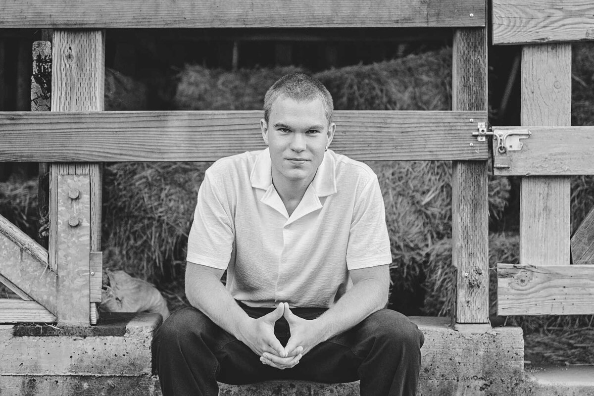 Black and white photo of a senior guy wearing a white shirt and black denim seated in a barn