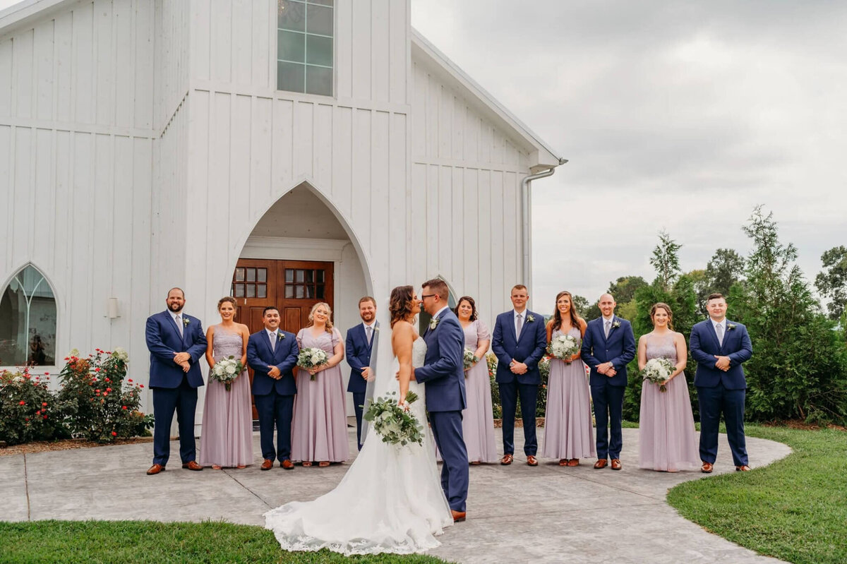 Photo of a bride and groom hugging while their wedding party stands in the background with a white wedding chapel