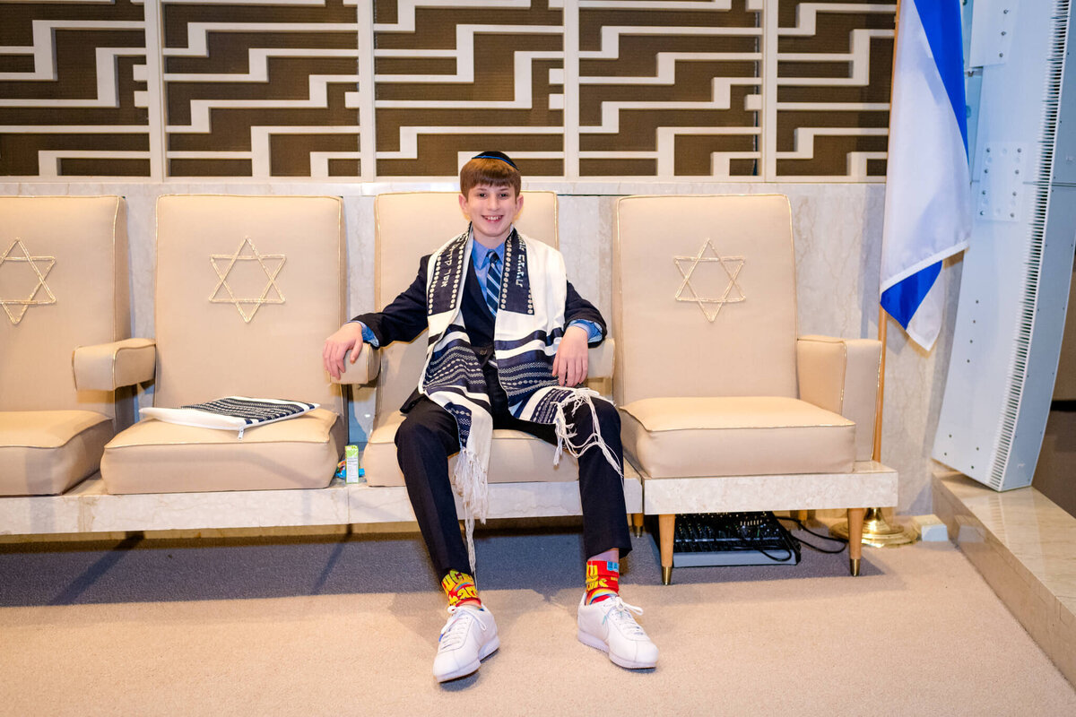 A boy in a blue suit and colorful socks sits in his tallit and kippah