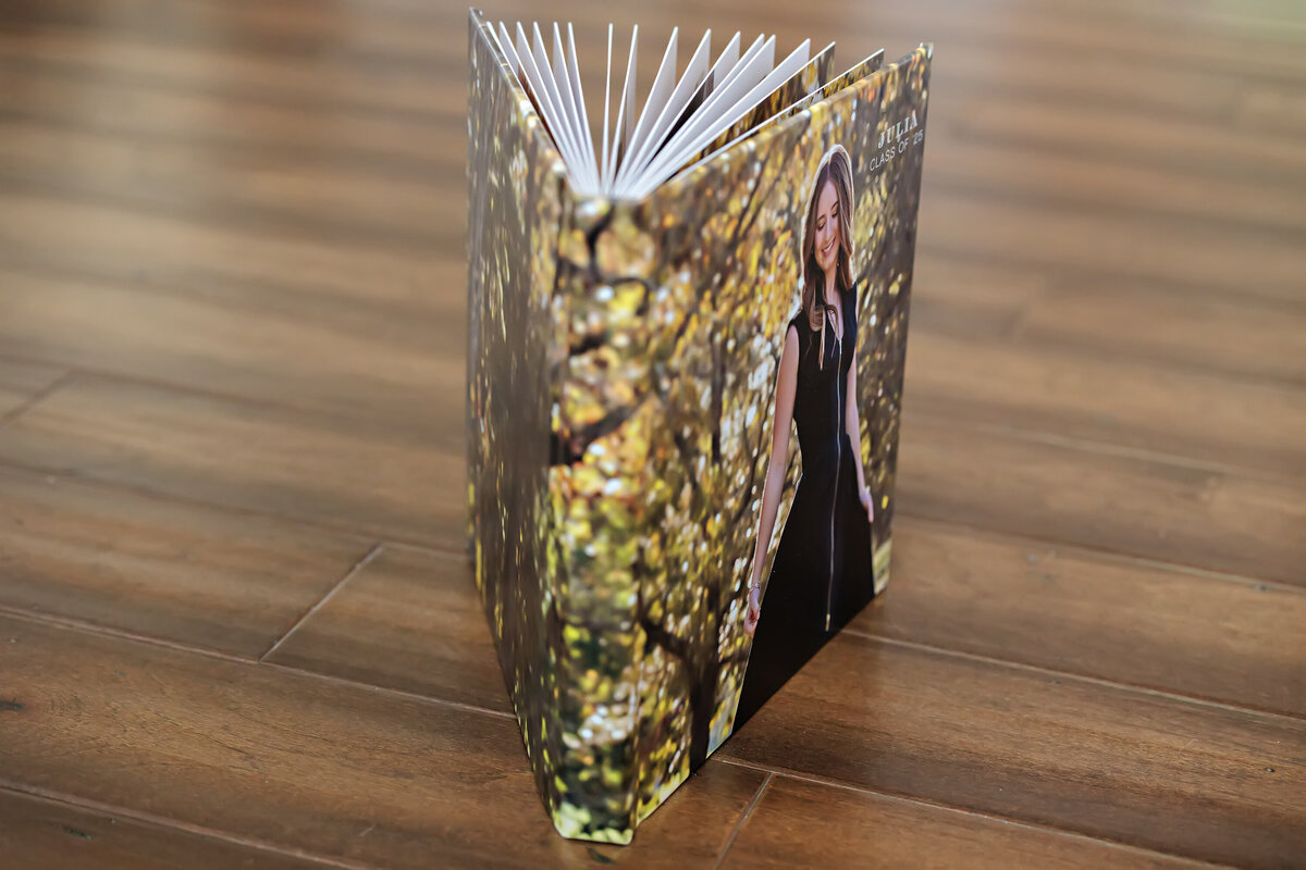 Photo Cover Session Book featuring a full wraparound image of a high school senior walking through vibrant fall foliage, shown standing upright with pages fanned to highlight thickness and premium print quality.