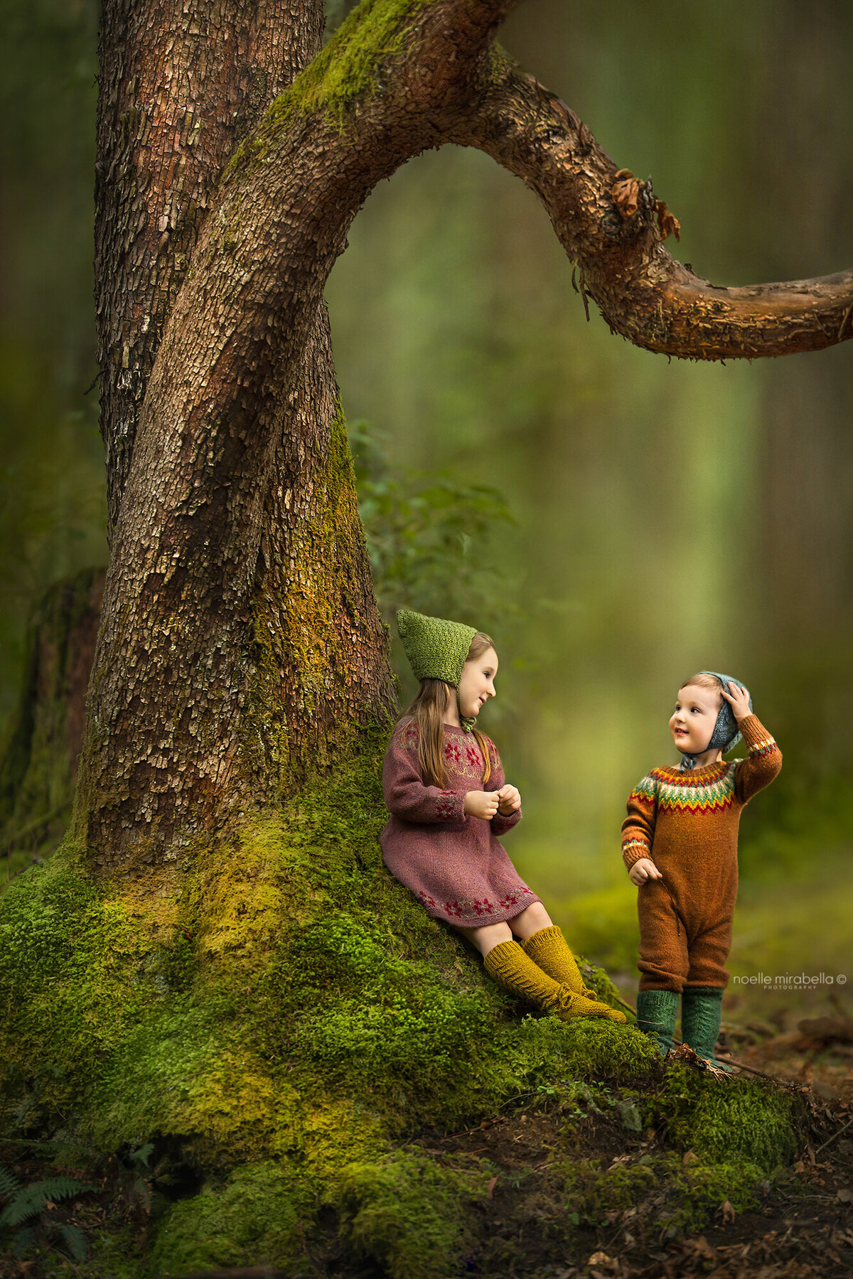 Children playing at the base of a large mossy tree in the forest, looking at each other laughing.