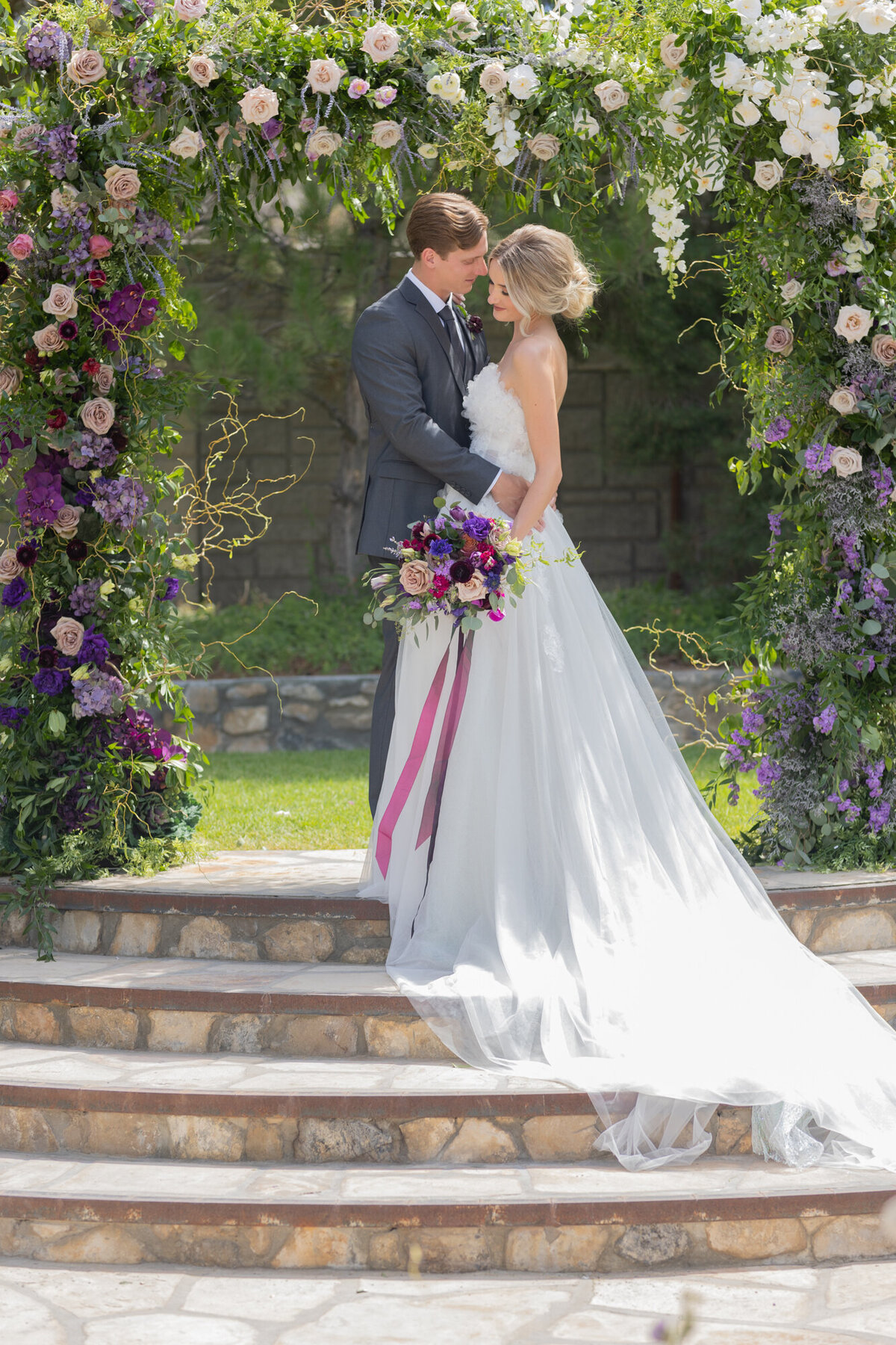 Bride and groom stand on steps under an arch of flowers