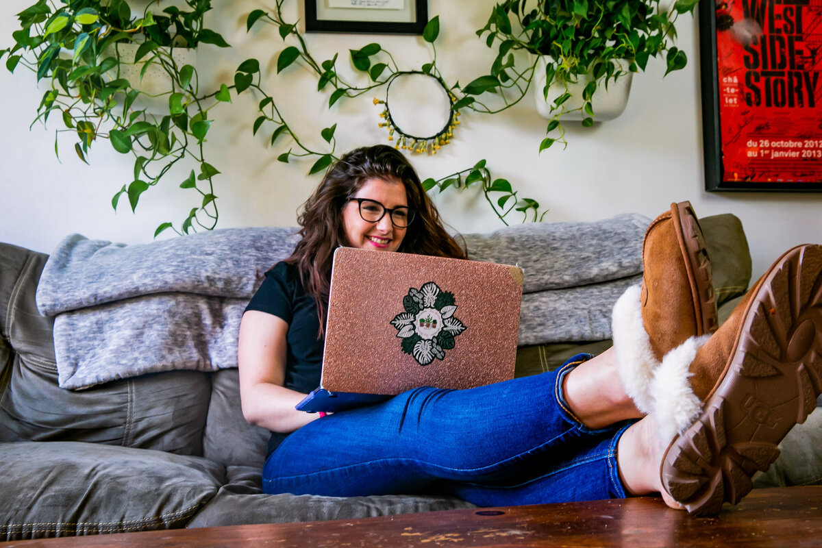 Host of Bloom & Grow Radio podcast, Maria,  sits on her sofa on her laptop surrounded by plants covering her wall