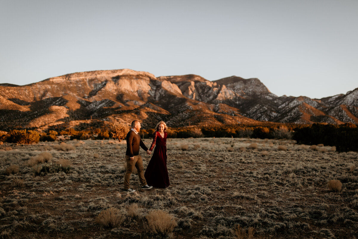 engaged couple walking through the desert with Sandia mountains behind them