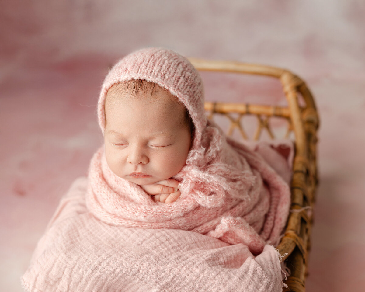 Newborn baby girl swaddled in a soft pink knit outfit with a matching bonnet, peacefully resting in a woven basket, surrounded by pink textures. Captured by Inland Empire newborn photographer Kaitlyn Dawn Photography.