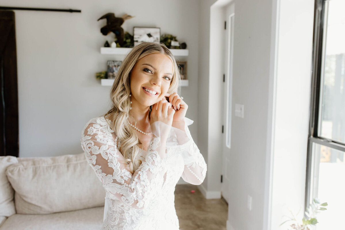 Bride putting on wedding day jewelry before wedding ceremony while wearing white lace wedding gown