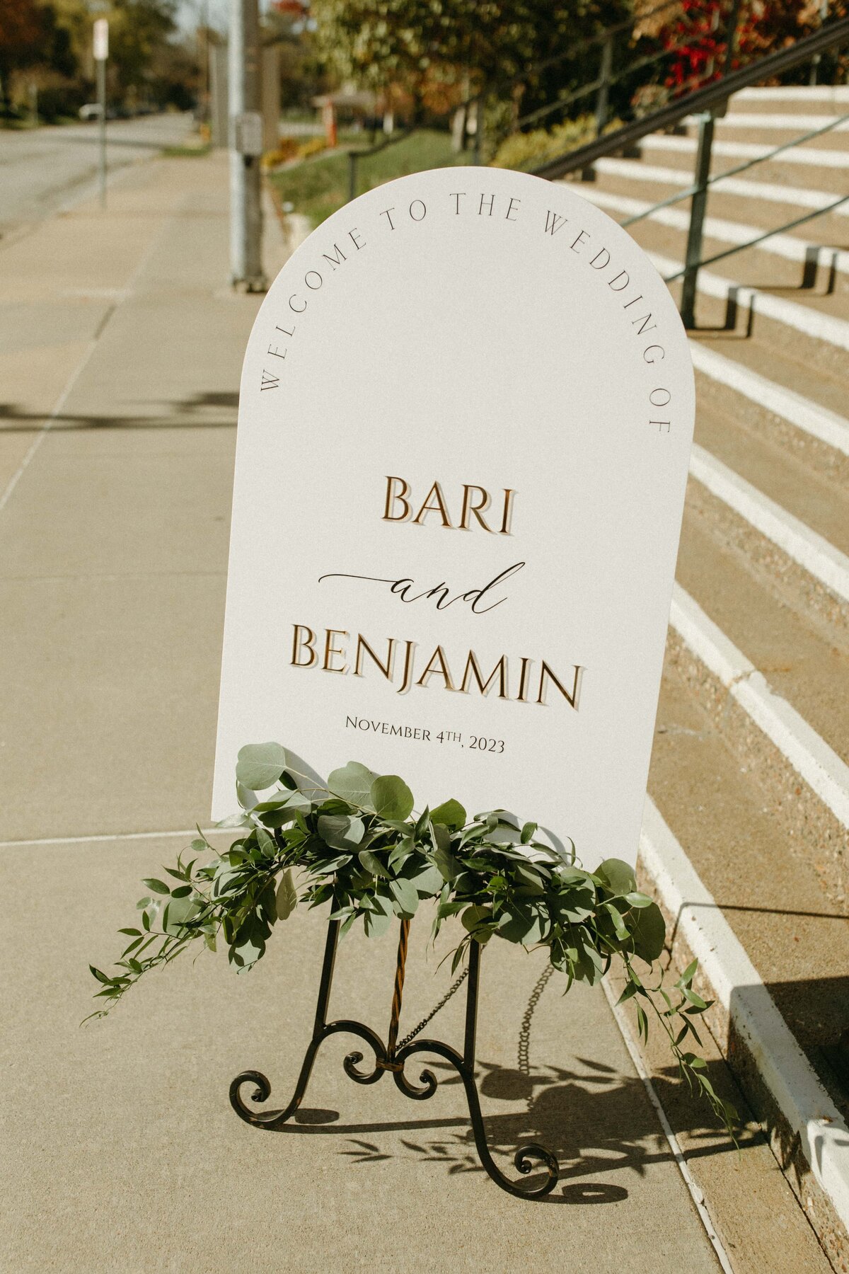 An elegant wedding welcome sign with the names "Bari and Benjamin" and the date "November 4th, 2023" on a sunny sidewalk in Davenport, adorned with green leaves.