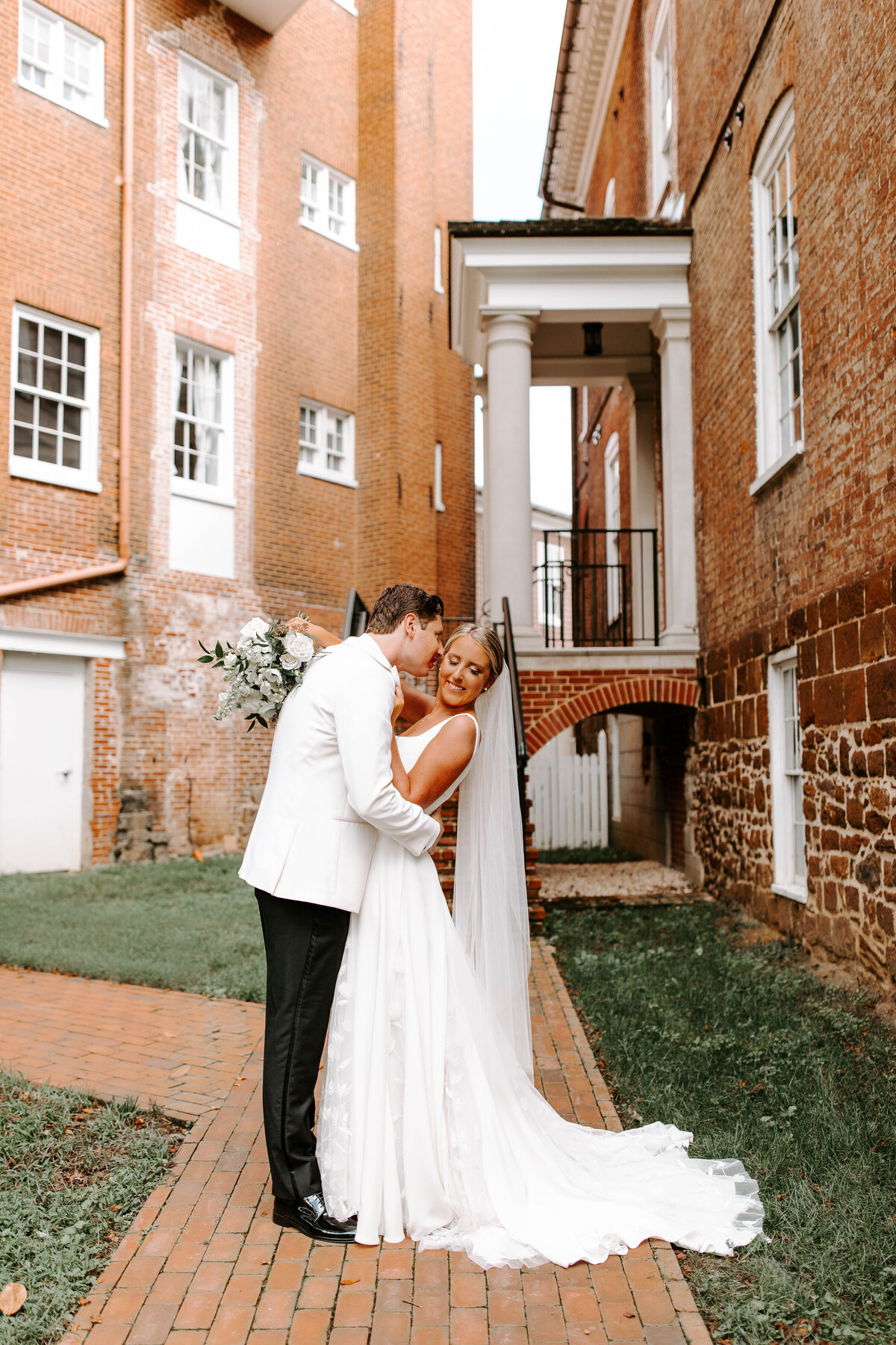 Bride and groom on their wedding day. The couple is standing stomach to stomach and  the groom has his nose on the side of the brides face as she smiles down towards her shoulder. The couple stands in front of a beautiful brick building.