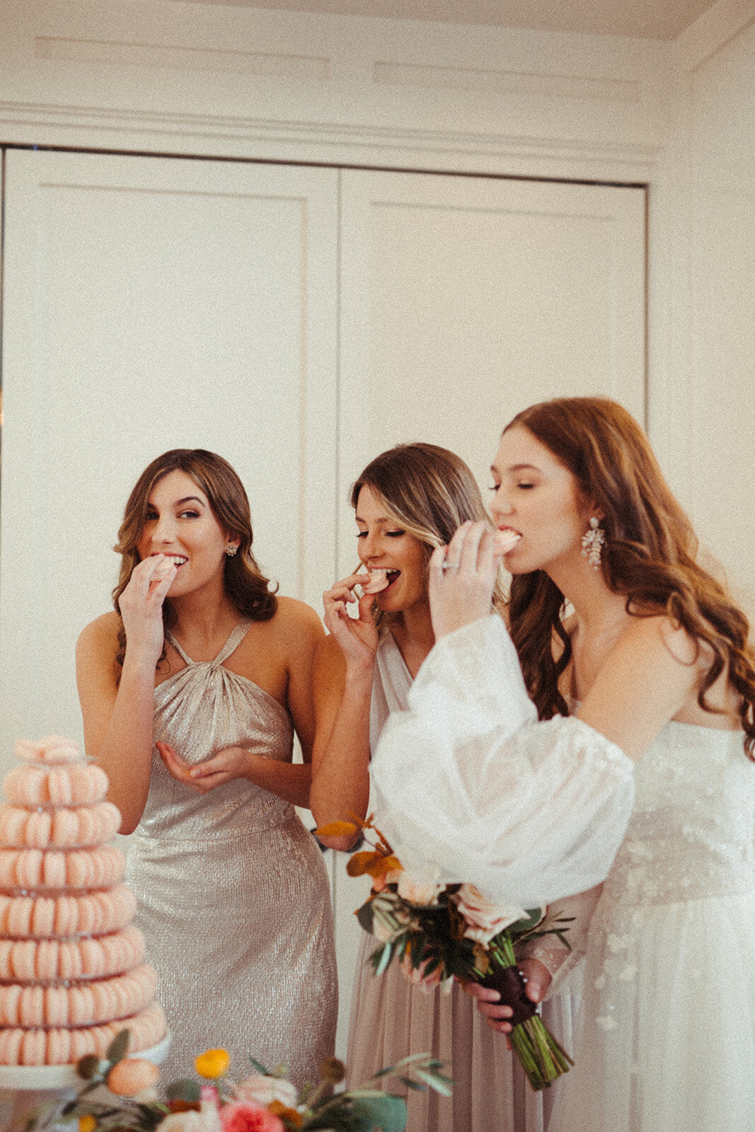 A bride wearing a white wedding gown and two women in dresses enjoy a bite of pastries beside a tower of peach-colored macaroons.