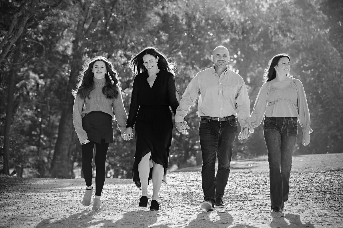 Monochrome image of parents and two daughters smiling while walking down tree‑lined gravel road at Anne Springs Close Greenway