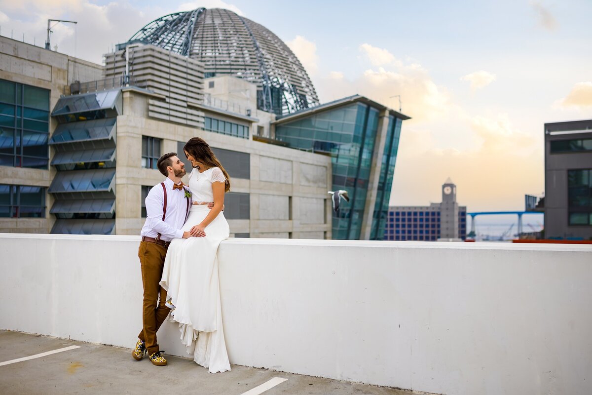 Bride and groom posing along wall on top of parking structure with a view of Coronado bridge and the San Diego library behind them