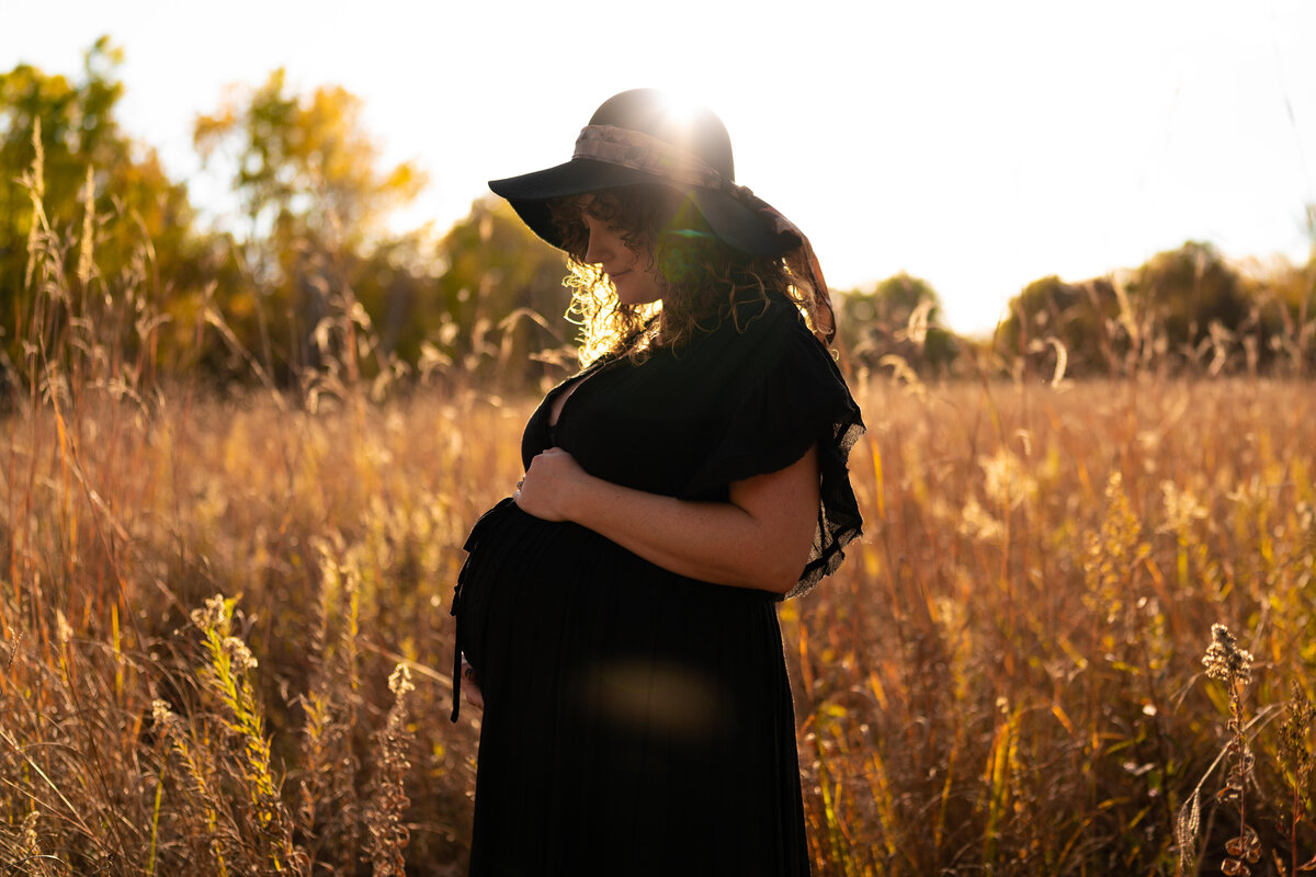 Pregnant woman in black dress and hat holds stomach in a field at sunset.