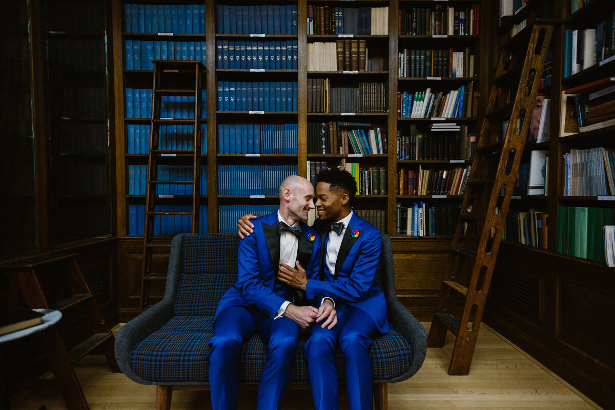 Two grooms sitting on a couch about to kiss.