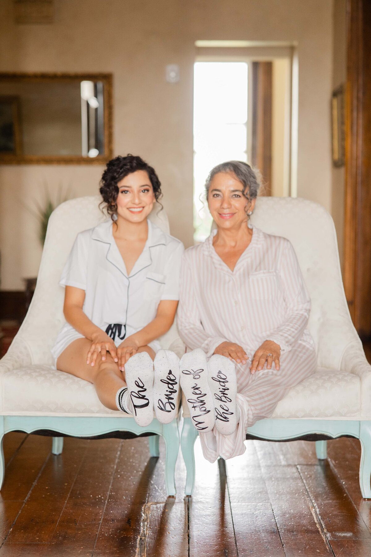 Two women in pajamas smiling on a sofa, displaying slippers that read "bride" and "mother of the bride," likely gearing up for an Iowa wedding.