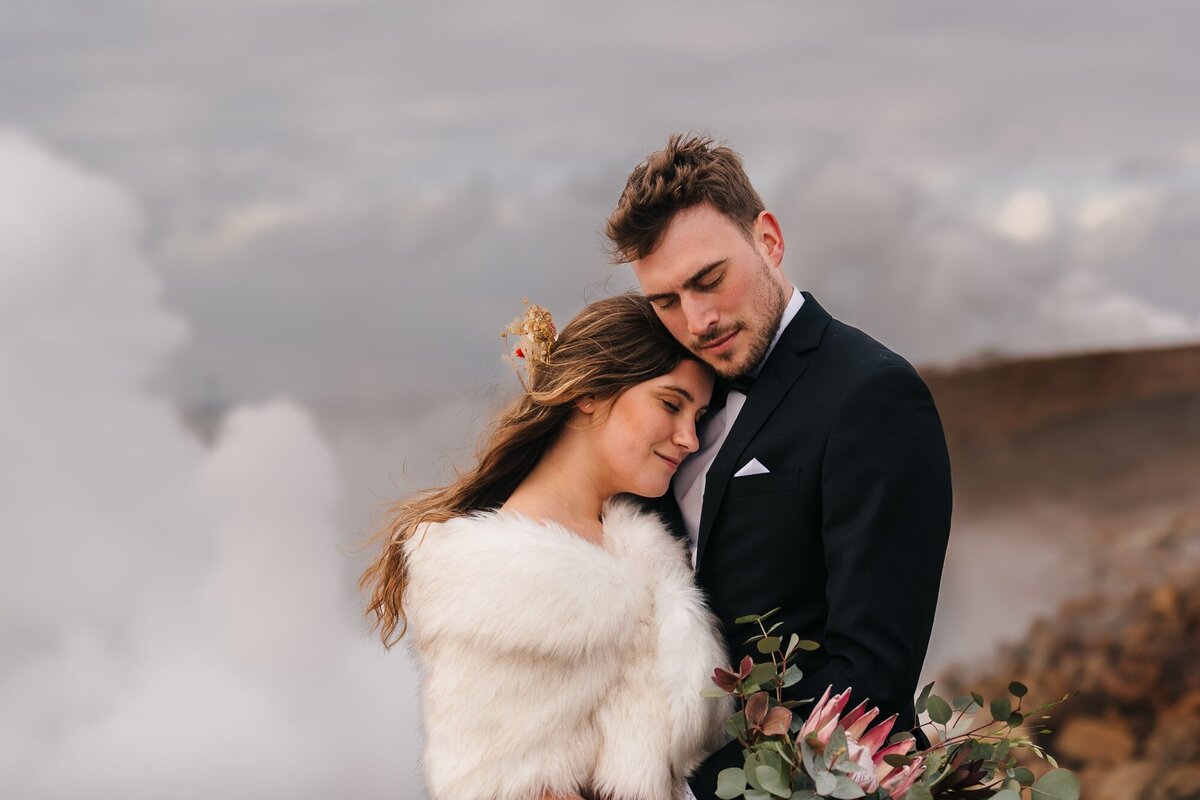 Amidst the geothermal wonders of Iceland, the bride rests her head on the groom's chest, sharing an intimate moment while the ethereal geothermal mist smoke around them.