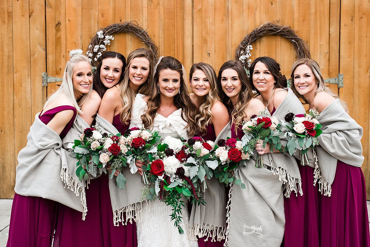 Bridesmaids surrounding bride wearing maroon dresses and shawls with red and white bouquets standing in front of a barn