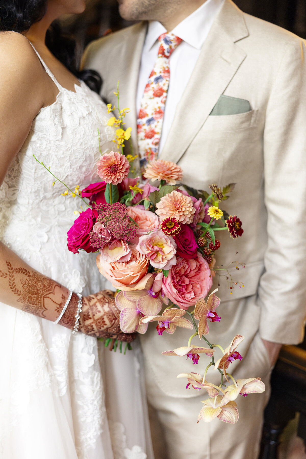 A couple stands close, wearing a white wedding dress and a beige suit. The bride holds a vibrant bouquet of pink, red, and peach flowers. Intricate henna designs adorn her hand, and the grooms floral tie complements the bouquet.