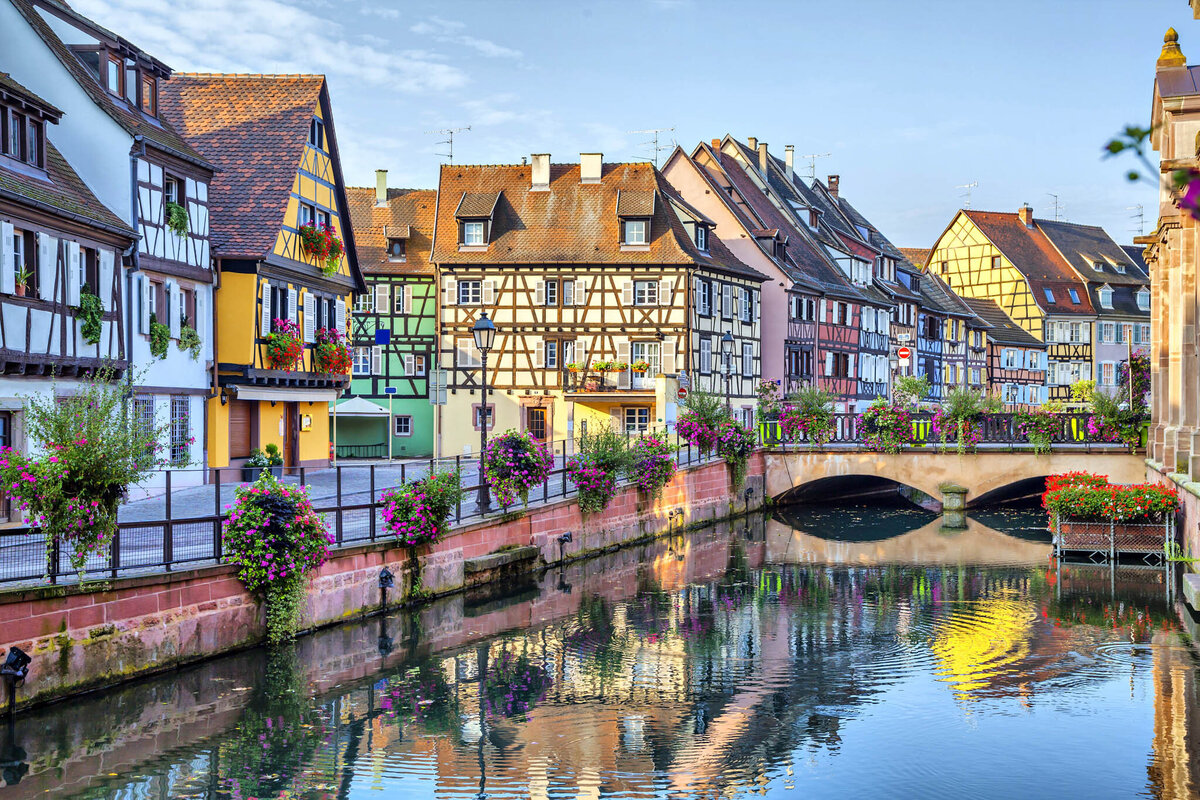 Colorful, traditional half-timbered houses line a canal in a picturesque town. Flower baskets adorn the railings, and a stone bridge arches over the water, reflecting the vibrant facade of the buildings against a clear blue sky.