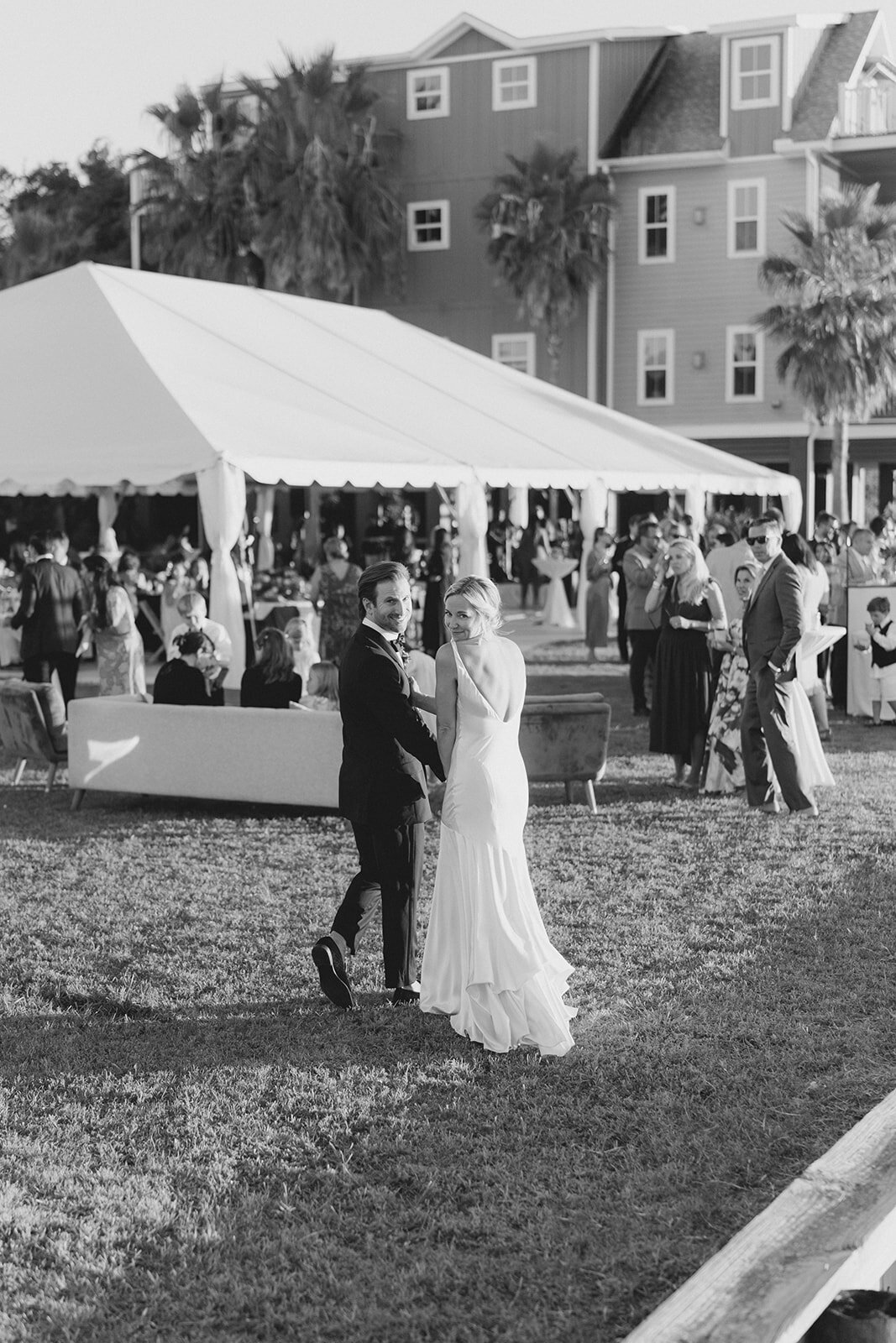 Waterfront fall wedding in Charleston. Bride and groom look back at the camera while walking to cocktail hour during sunset. Kailee DiMeglio Photography. Black and white wedding photographer.
