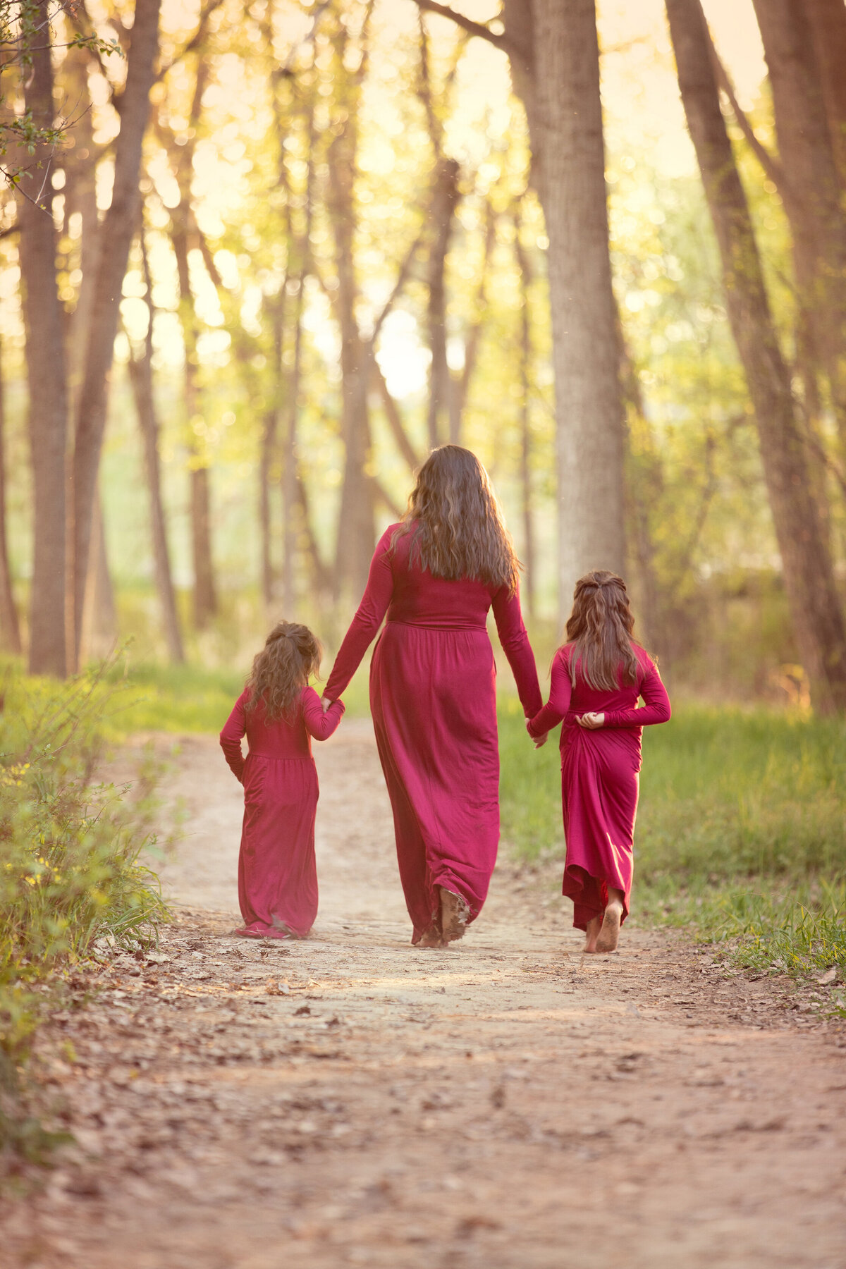 Mom and daughters wearing maroon dresses walking along path at river front park near the river at sunset.