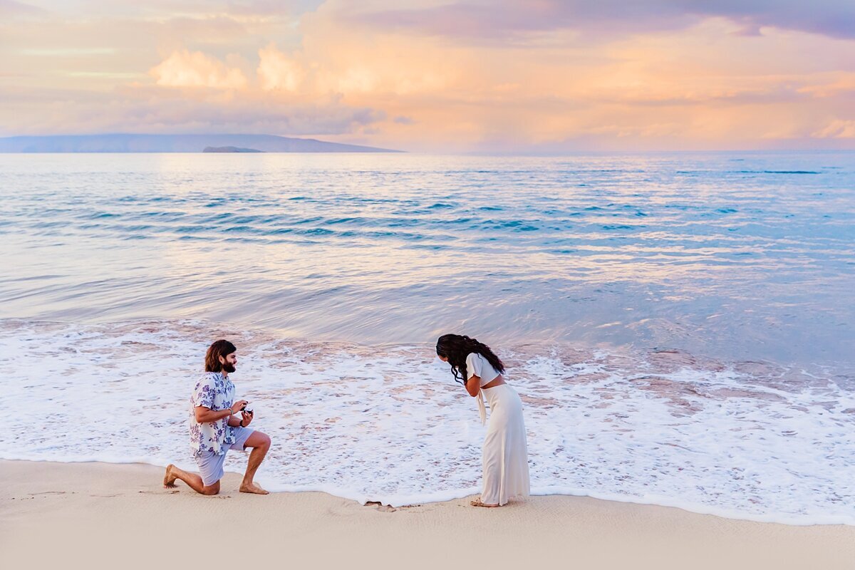 Brunette man gets down on one knee to propose to his partner on a beach at sunset in Maui
