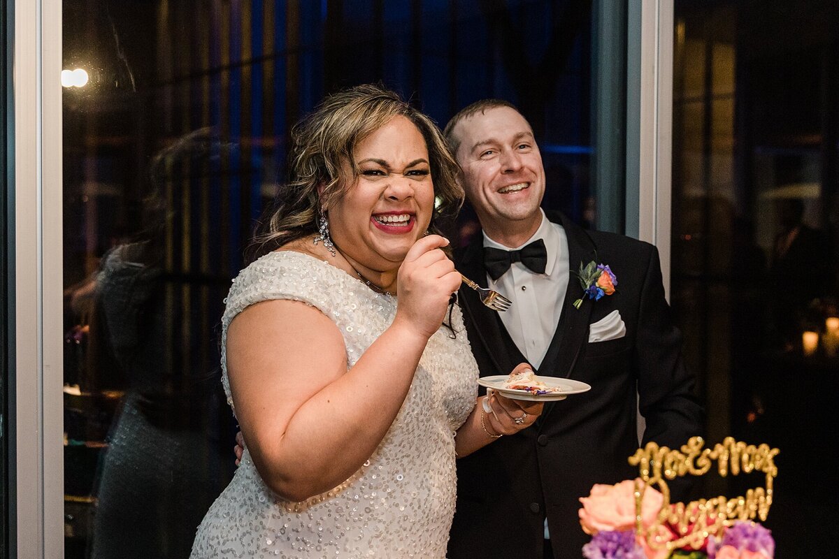 A candid moment of a bride and groom laughing after their caking cutting during their wedding reception at the Modern Art Museum of Fort Worth in Fort Worth, Texas. The bride is on the left and is wearing a highly detailed, white dress and is holding a plate of cake in one hand and a fork in the other. The groom is on the right and is wearing a black tuxedo, bowtie, and boutonniere. The top of their cake can be seen covered with flowers and a gold cake topper with their last name on it.