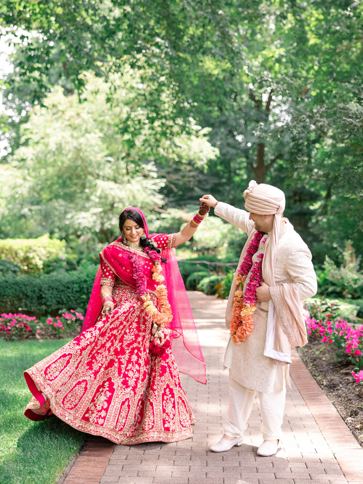 A couple dances on a garden path. The woman wears a vibrant pink and gold traditional dress, while the man wears a cream outfit with a turban. Both are adorned with floral garlands. The background is lush with greenery and flowers.