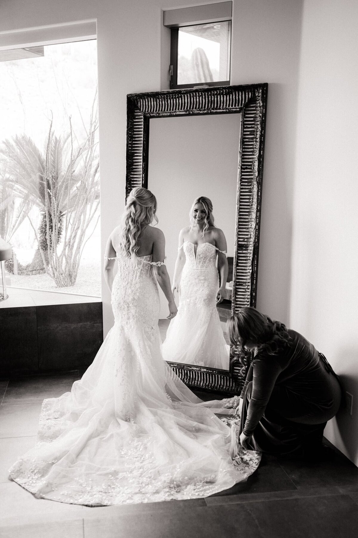 Bride looking at herself in a large mirror while bridesmaid fixes her train