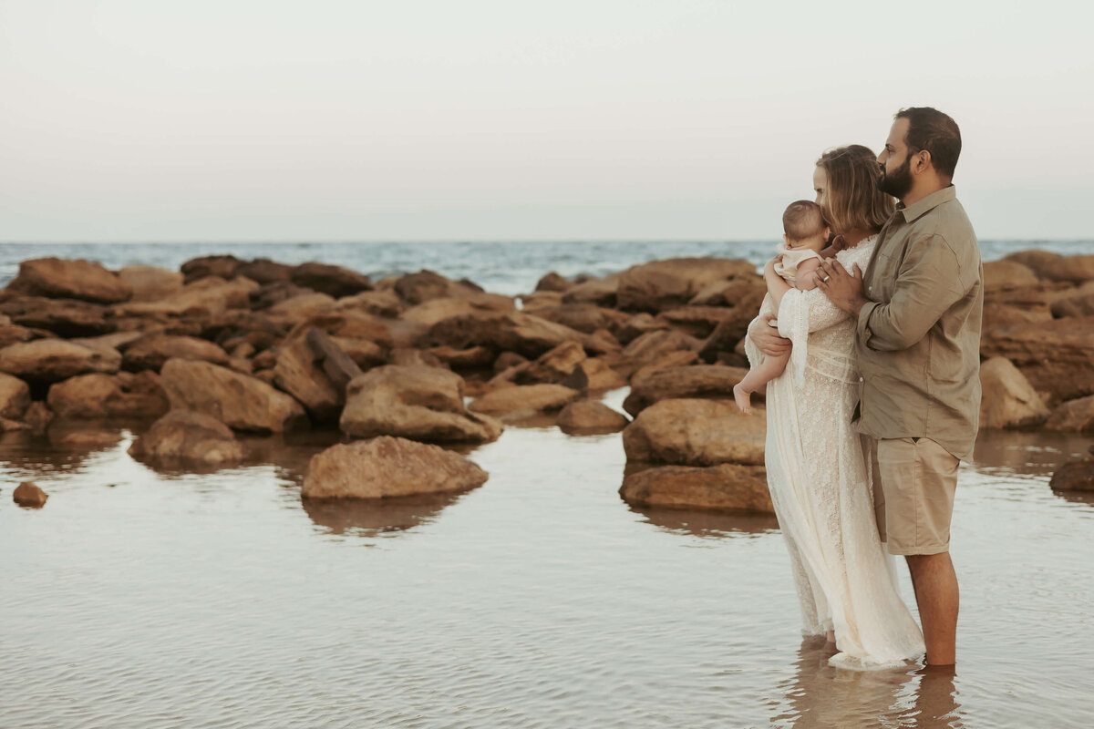 Parents and their baby standing close with the sea and rocks behind them.