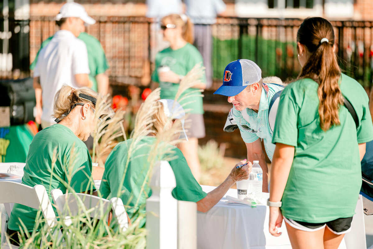 A group of people gathered outdoors by a registration table. Two women in green shirts sit and interact with a man in a blue hat and striped shirt. An event photographer from Fayetteville, NC captures the scene as a young woman in green stands nearby, with others in casual attire visible in the background.
