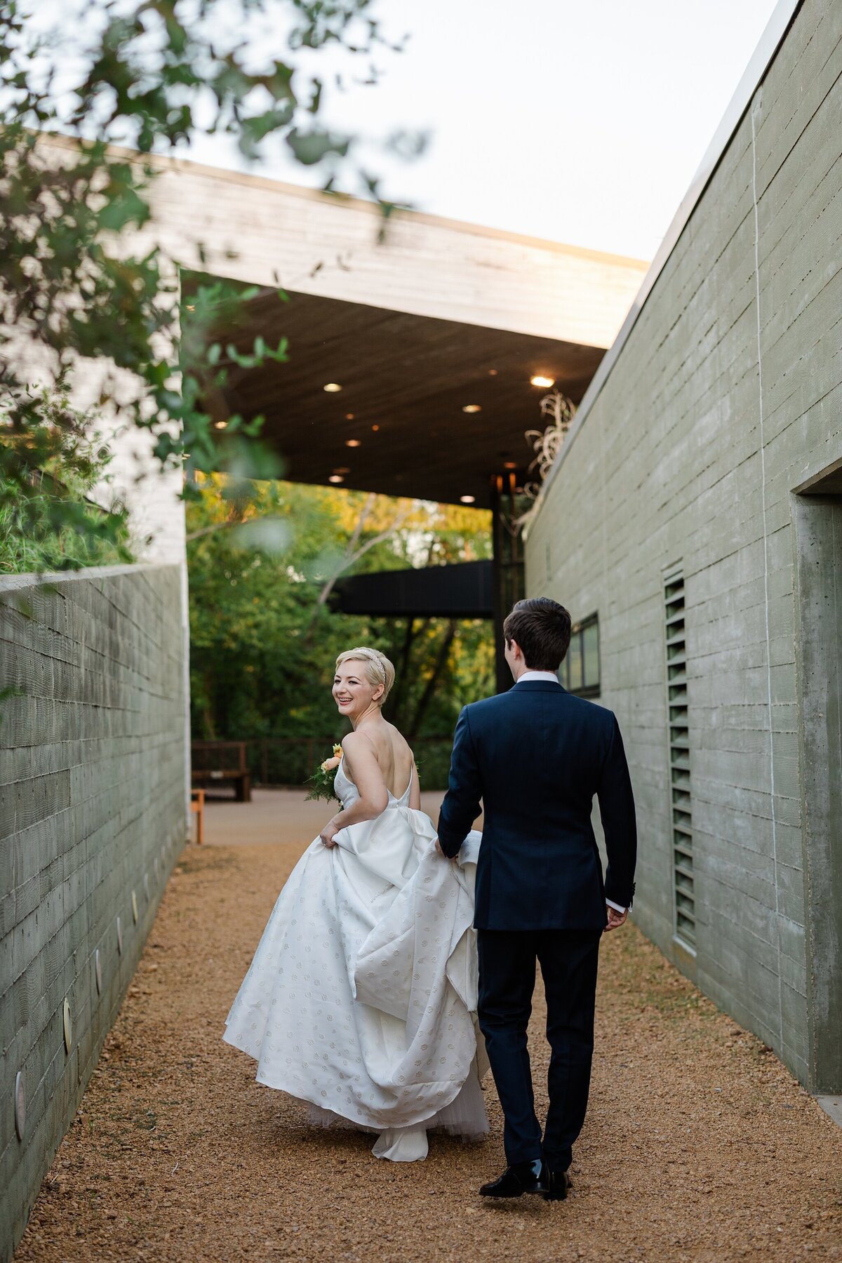 A portrait of a bride and groom walking away from the camera on their wedding day at the Trinity River Audubon Center in Dallas, Texas. The bride is on the left and is looking back at the camera. She wears a long, flowing, short sleeve white dress. The groom is on the right and is wearing a navy suit.