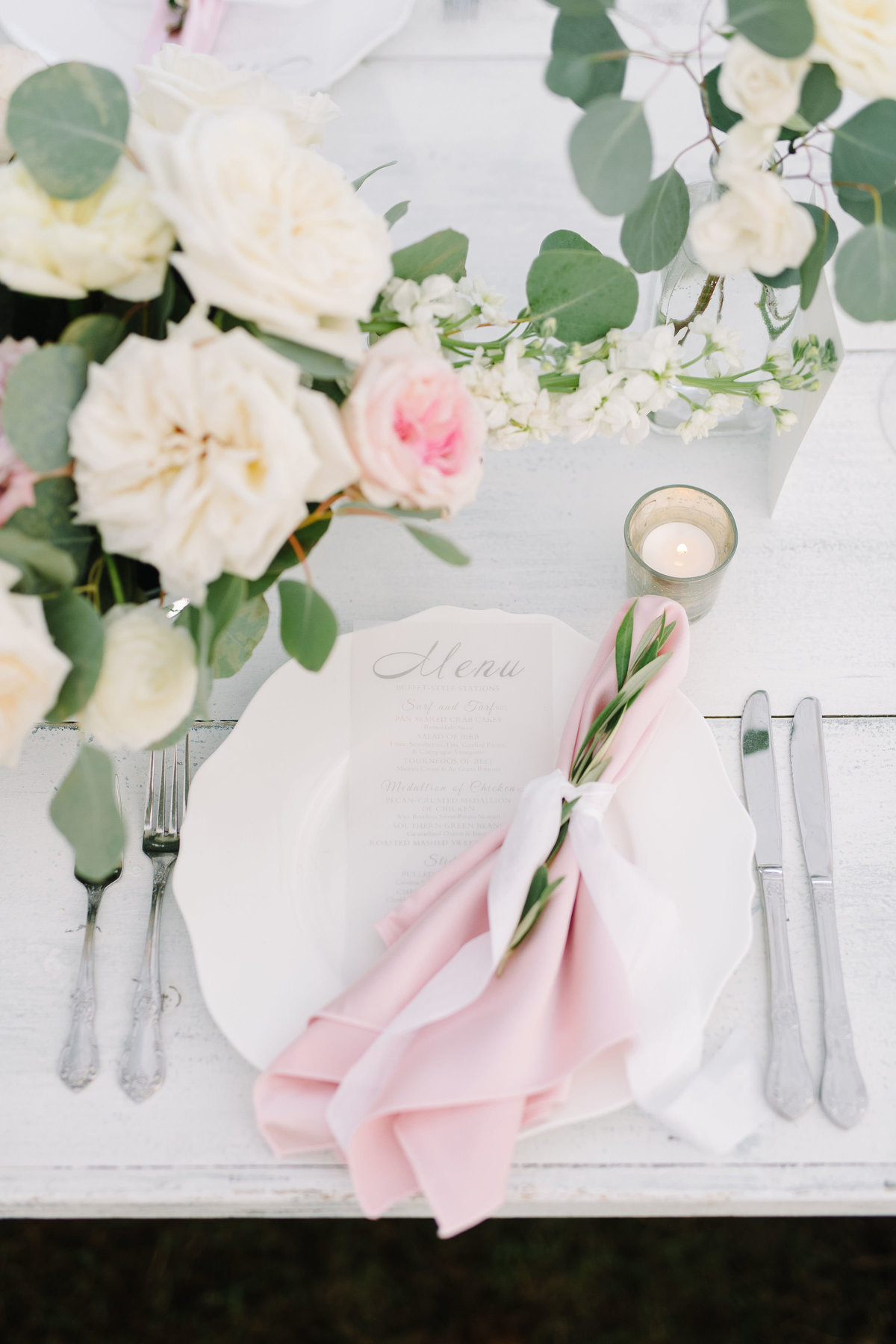 Blush Pink Napkin Fold tied with Ribbon and Sprig of greenery on White Farm Table with Silverware Place Setting
