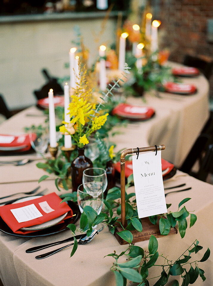 Curved table set with ivory table cloth, red napkins, light candlesticks, and amber glassware with flowers.