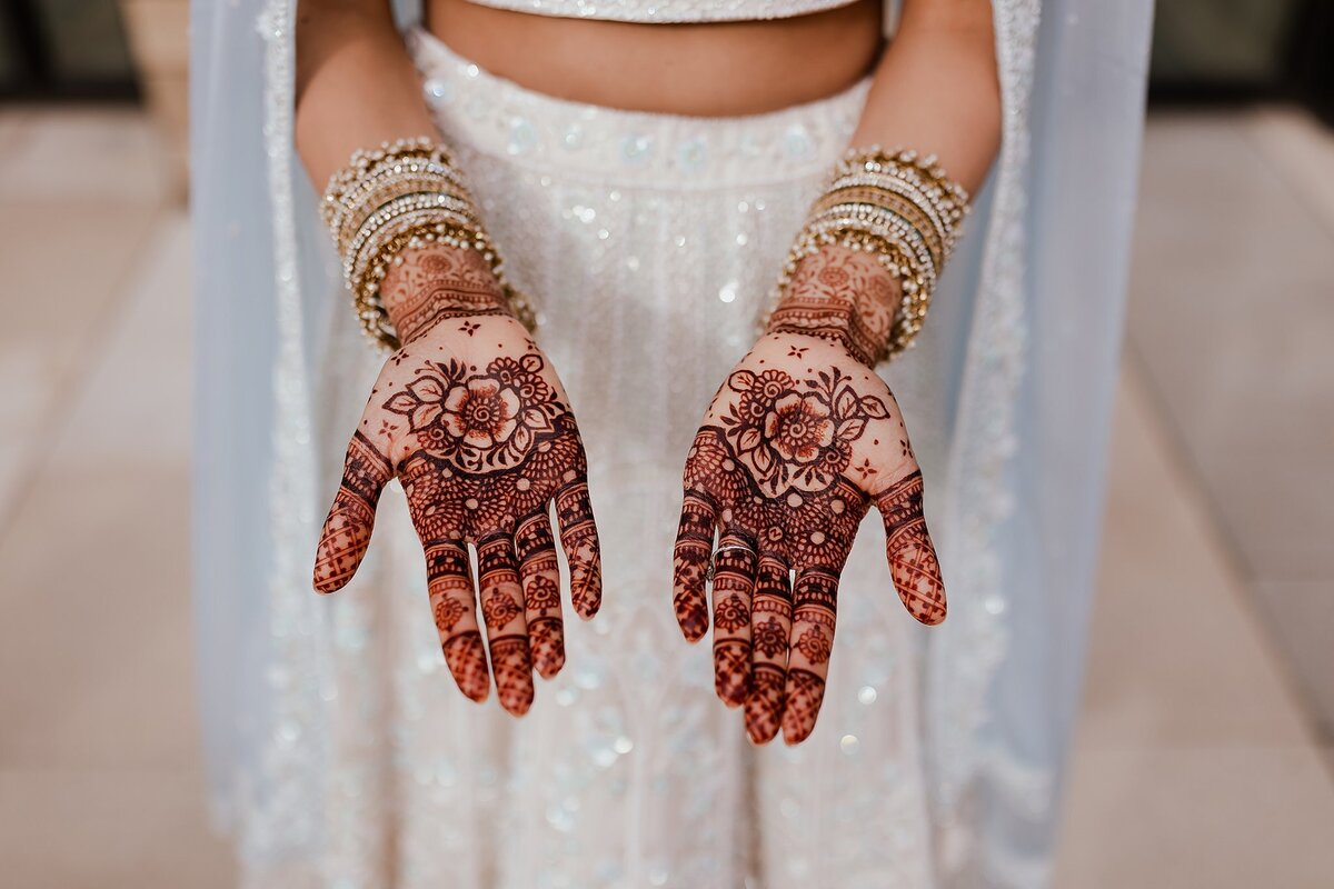Indian bride shows off the henna on her hands from her wedding mendhi. The hindu bride is wearing a white beaded saree with a light blue dupatta and white and gold Indian wedding jewelry.