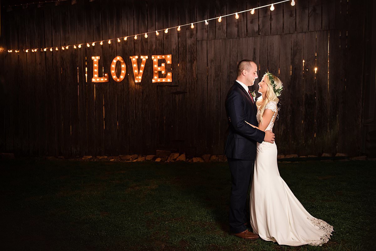 Bride and groom snuggled together at night with the love light up letters on the outside of Drakewood Farm barn