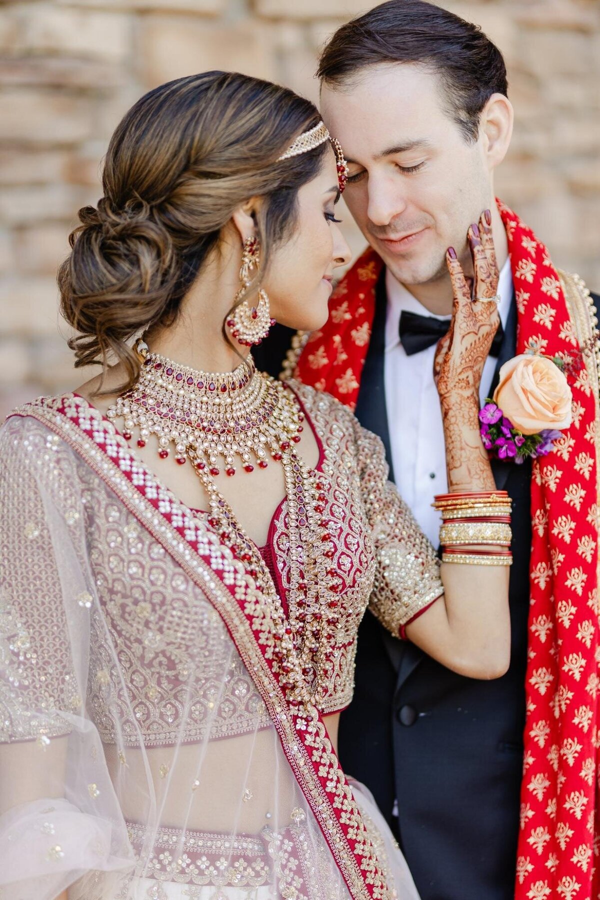 Bride in detailed traditional indian attire and groom in a black tuxedo with a red scarf, tenderly touching faces.