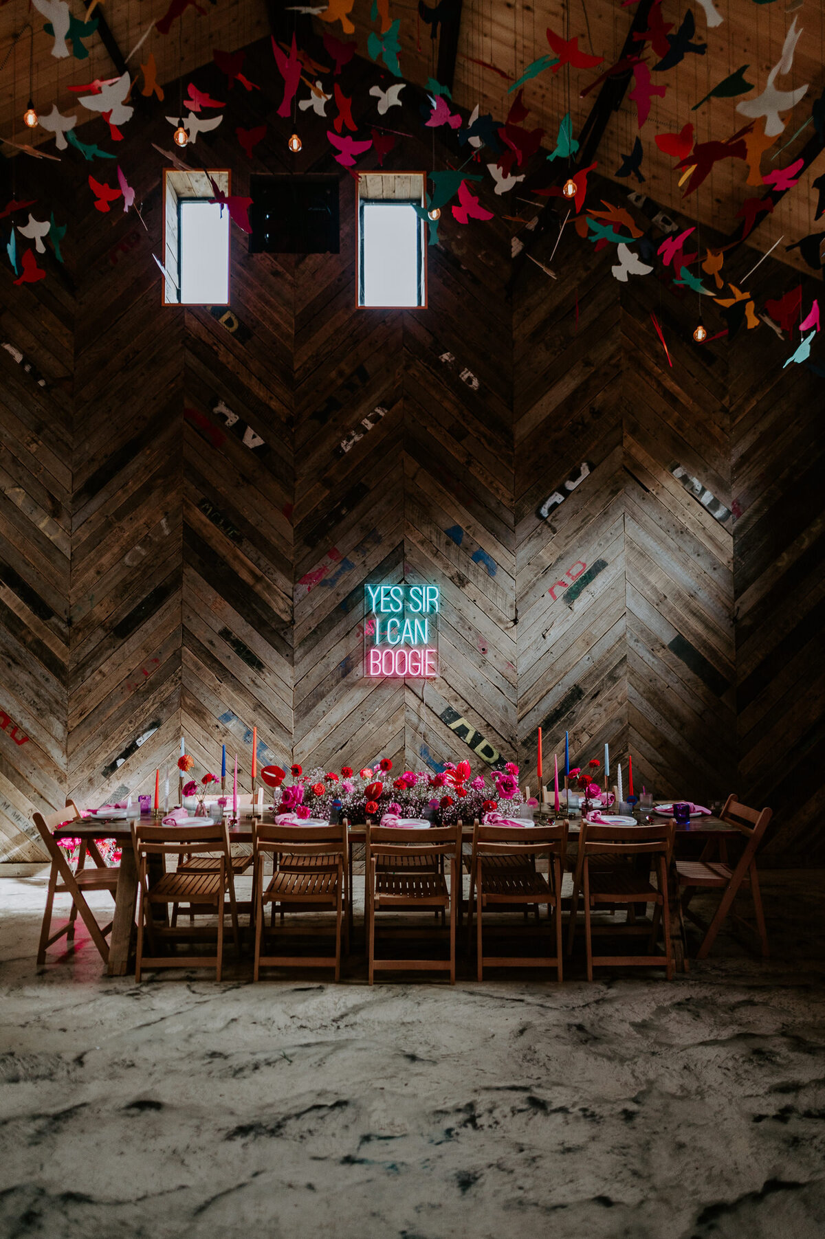 Pink wedding styling at The Canary Shed. A pink and blue neon which reads Yes Sir I Can Boogie hangs above a large dressed table. The disco themed wedding has a strong pink and purple theme.