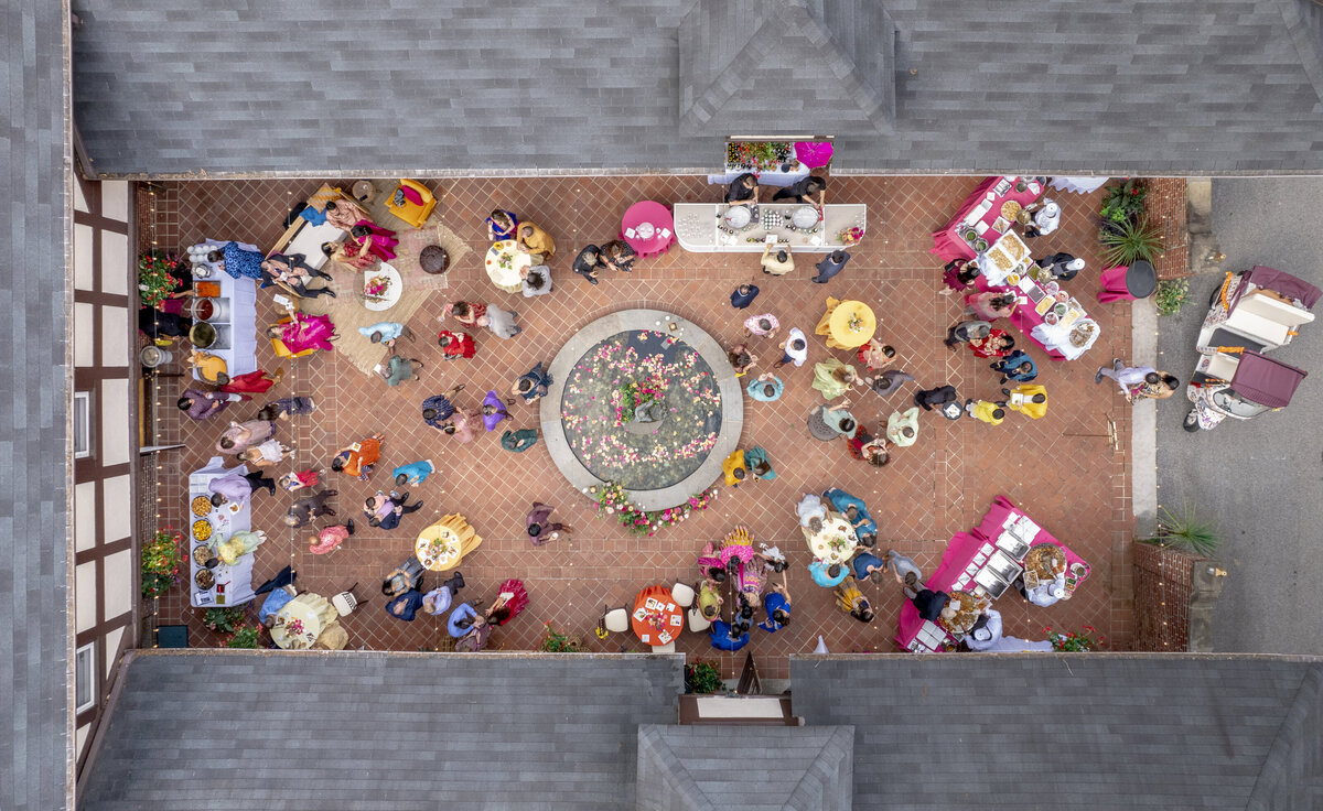 Aerial view of a vibrant outdoor gathering in a courtyard. Guests in colorful attire sit at round tables, surrounded by festive decorations and food stands. A central floral display adds a focal point to the brick-patterned flooring.