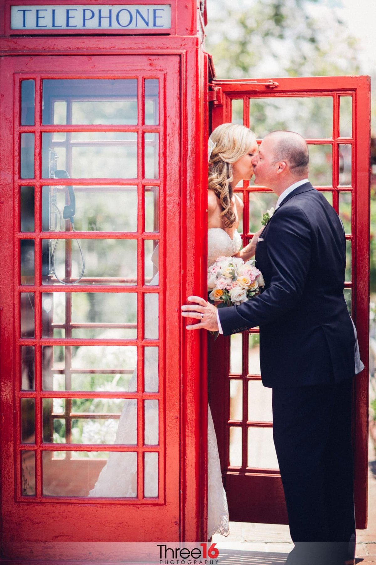 Bride stands in an older looking English telephone booth as leans out to kiss her Groom