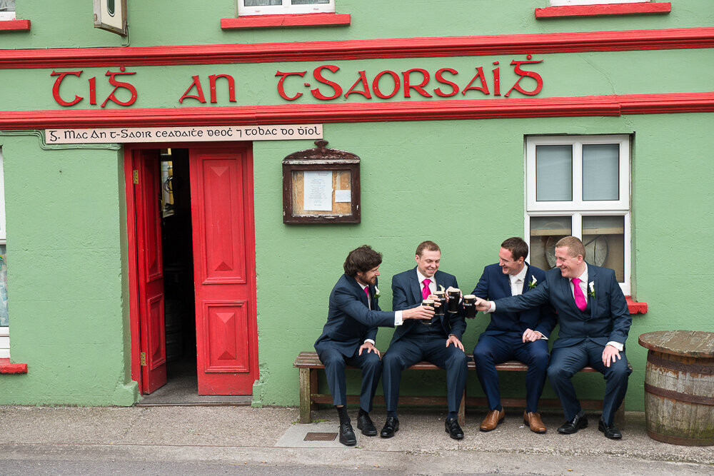 Groom and groomsmen wearing navy suits and pink ties, sitting outside a green and red painted, traditional Irish pub in Ballyferriter, Kerry
