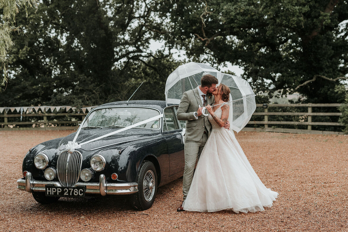 Bride & Groom kiss under clear umbrellas in front of their vintage wedding car at their barn wedding at Odo's Barn, Ashford